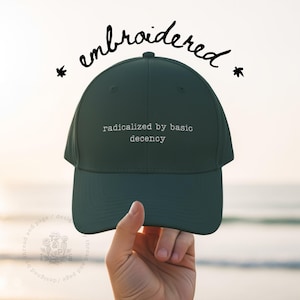 Op de afbeelding: Donkergroene baseballcap met de witte tekst "radicalized by basic decency". Het woord "embroidered" staat in cursief boven de pet. De pet wordt tegen een vage achtergrond gehouden.
