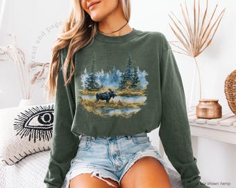 May include: Olive green long-sleeve sweatshirt featuring a nature scene with a moose, trees, and water. The graphic is in a circular design. The sweatshirt is made of a soft material. The text "read and page" is visible.