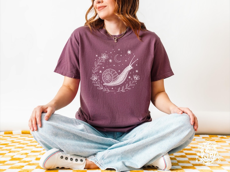 May include: A purple t-shirt with a white graphic of a snail with a shell, surrounded by flowers and stars. The snail is crawling up a vine. The t-shirt is on a person sitting with their legs crossed.
