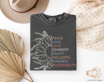 May include: Dark gray t-shirt featuring a white sunflower design on the left side and the words "PEACE, HOPE, LOVE, DIVERSITY, EQUITY, INCLUSION, KINDNESS, ACCESSIBILITY" in various colors on the right. A tan hat and decorative fan are in the background.
