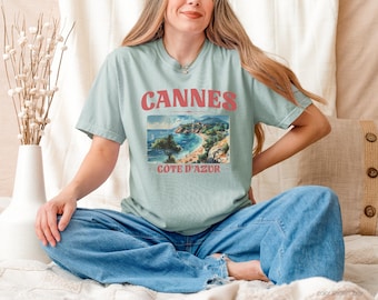 May include: A light blue t-shirt with the word "Cannes" in red letters and a scenic image of the French Riviera. The shirt also has the words "Cote D'Azur" in red. The person is wearing blue jeans.