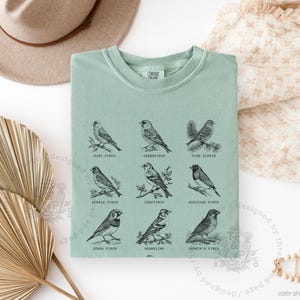 May include: Sage green t-shirt featuring nine detailed illustrations of different bird species, each labeled with their common name. The design is in black ink, creating a vintage aesthetic. The shirt is folded neatly on a white surface.
