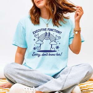 Puede incluir: Camiseta azul claro con el texto "EXECUTIVE FUNCTION? Sorry, don't know her!" y un diseño de arcoíris de dibujos animados. La camiseta es de color chambray.
