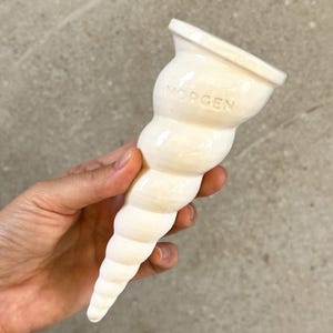 May include: A cream-colored, ceramic seashell-shaped object with the word "MORGEN" inscribed on the top. The object has a flared opening and a spiraling, textured body. The piece is held in a hand against a neutral background.