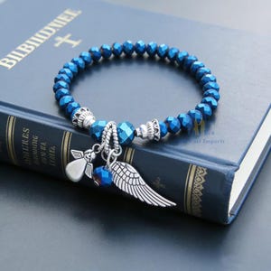 Puede incluir: Una pulsera el&aacute;stica compuesta por cuentas facetadas de color azul met&aacute;lico. La pulsera presenta tres dijes de tono plateado: una peque&ntilde;a figura de &aacute;ngel, un ala individual y una cuenta azul peque&ntilde;a. Los dijes est&aacute;n unidos por un anillo met&aacute;lico.