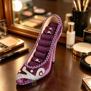 May include: A purple velvet high-heeled shoe-shaped ring holder. The interior features multiple slots for rings, lined with a deep purple fabric. The shoe is decorated with silver glitter accents and white trim. The heel is a matching purple color.
