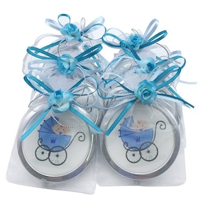 May include: Baby shower favors featuring a blue baby carriage design. Each favor is a round compact mirror, packaged in a clear bag and adorned with blue ribbons and a decorative blue rose. The favors are ideal for a baby boy.