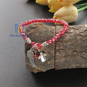 Puede incluir: Una pulsera el&aacute;stica con cuentas de cristal rojo facetado y dos dijes de metal plateado, uno con forma de &aacute;ngel y otro con forma de ala. La pulsera incluye peque&ntilde;as cuentas espaciadoras plateadas y el texto West East Imports.