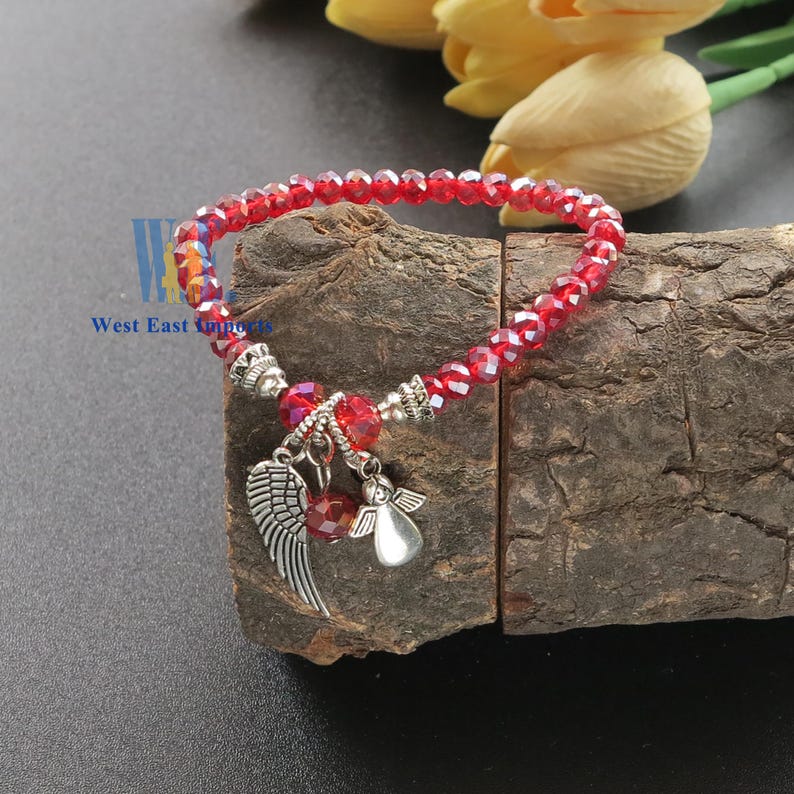 Puede incluir: Una pulsera el&aacute;stica con cuentas de cristal rojo facetado y dos dijes de metal plateado, uno con forma de &aacute;ngel y otro con forma de ala. La pulsera incluye peque&ntilde;as cuentas espaciadoras plateadas y el texto West East Imports.