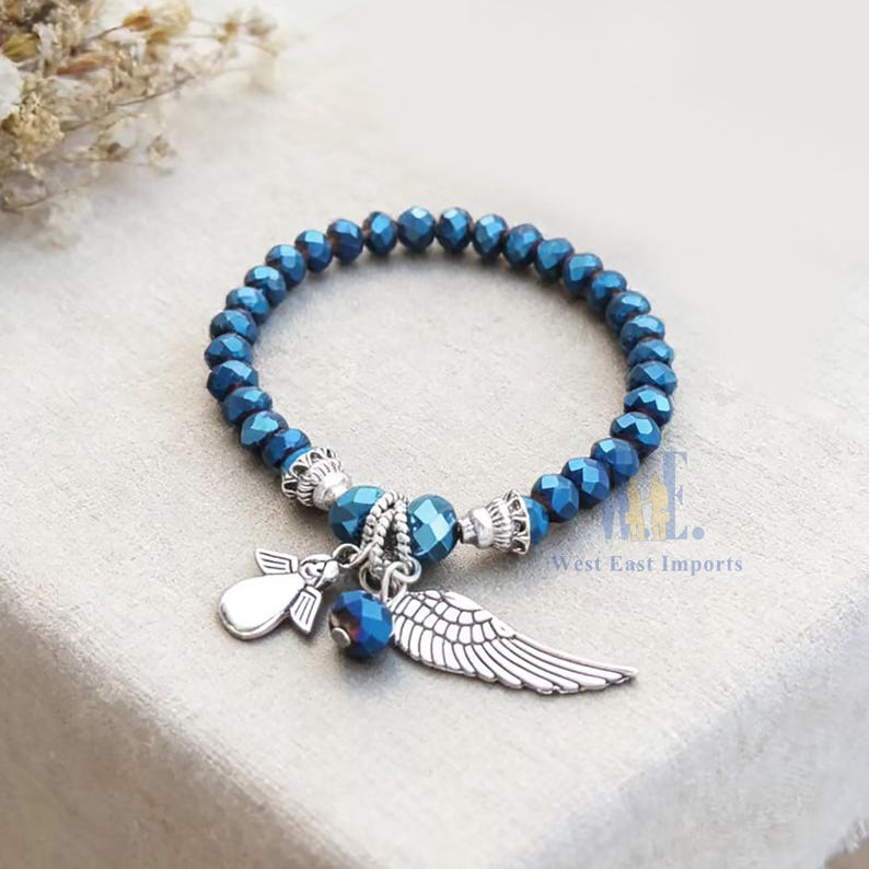 Puede incluir: Una pulsera el&aacute;stica con cuentas azul oscuro facetadas y detalles en metal plateado. La pulsera incluye tres dijes colgantes: una peque&ntilde;a figura de &aacute;ngel plateada, una cuenta azul y un dije de ala plateada detallado. El texto dice West East Imports.