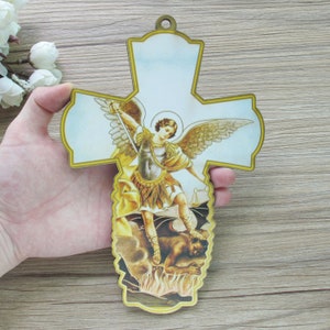 May include: A wooden cross with a gold trim features a colorful image of an angel with a sword defeating a demon. The angel is depicted with wings and a halo, while the demon is shown with horns and a tail.
