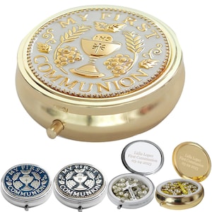 May include: A gold-colored pill box with the words "MY FIRST COMMUNION" in a circular design. The design includes a chalice, grapes, and wheat stalks. Additional pill boxes in blue and silver are also shown.