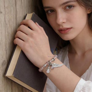 Puede incluir: Una pulsera de cuentas con cuentas esf&eacute;ricas de color rosa claro transl&uacute;cido, espaciadores de tono plateado y dos dijes de metal colgantes. Un dije tiene forma de figura de &aacute;ngel estilizada y el otro tiene forma de ala emplumada.