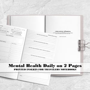 May include: A set of white paper inserts for a traveler's notebook featuring black printed text and lines for mental health tracking. The pages include sections for daily intentions, meals, reflections, and self-care. A silver metal binder clip holds the pages together.