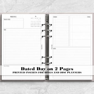 May include: A light gray six-ring binder planner open to two white pages with black text. The left page features a daily schedule, tasks, and a calendar. The right page includes sections for goals, fitness, and notes. A silver pen is attached to the side.