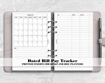 May include: A light gray ring-bound planner containing white paper inserts with a black ink printed January 2025 calendar on the left page and a bill payment tracker table on the right page, featuring a silver metal pen attached to the side.