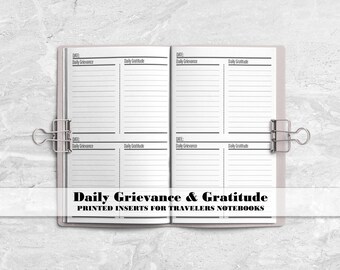 May include: An open notebook with white pages featuring black printed lines and headers labeled Date, Daily Grievance, and Daily Gratitude. Two silver metal binder clips are attached to the sides of the notebook, which displays the text Daily Grievance & Gratitude Printed Inserts for Travelers Notebooks.