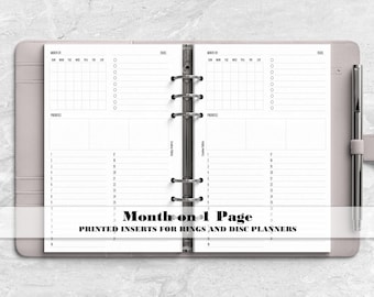 May include: A light gray six-ring binder planner open to two pages featuring a monthly calendar grid, a tasks list, a priorities section, and a numbered daily log. A silver pen is held in the side loop. Text reads Month on 1 Page.