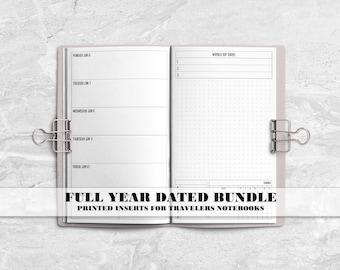 May include: An open planner with white pages featuring a weekly layout on the left and a dot grid section on the right, held open by two silver metal binder clips. The text reads FULL YEAR DATED BUNDLE PRINTED INSERTS FOR TRAVELERS NOTEBOOKS.