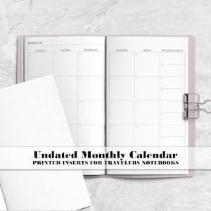 May include: Open undated monthly calendar insert for traveler's notebooks. The calendar pages are white with a grid layout, held together with a silver binder clip. The text "Undated Monthly Calendar" is displayed.