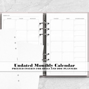May include: A light-colored ring binder planner containing undated monthly calendar pages with a grid layout, accompanied by loose dot grid paper inserts. A silver pen is attached to the side of the binder, which features the text Undated Monthly Calendar PRINTED INSERTS FOR RINGS AND DISC PLANNERS.