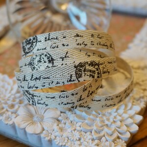 May include: Cream-coloured ribbon with a woven texture, featuring black script and stamp designs. The ribbon is coiled, showcasing its intricate details and gold accents. The text includes the words "York" and "June 7-PM".