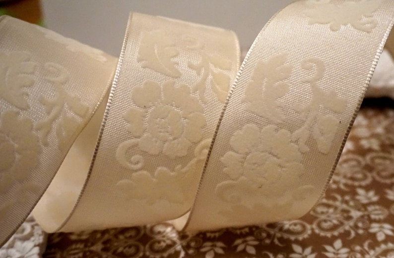 May include: White satin ribbon with an embossed floral design. The ribbon is curled and shows the design on both sides.