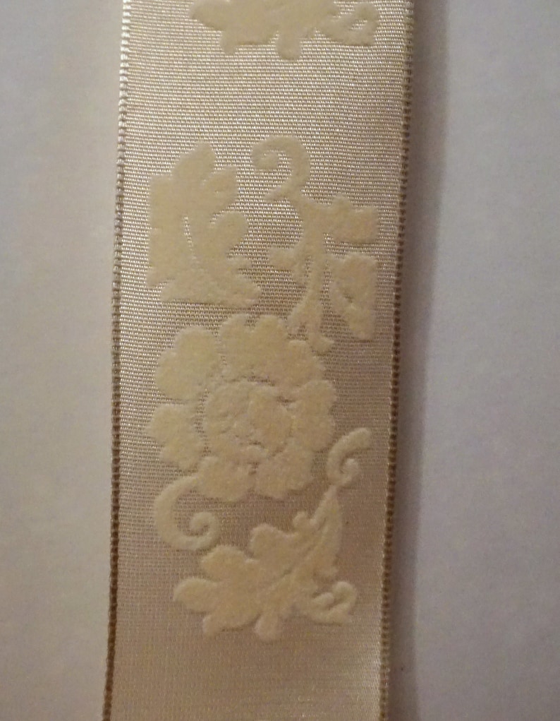May include: A close-up of a white satin ribbon with a raised floral design. The ribbon has a subtle sheen and a delicate texture.