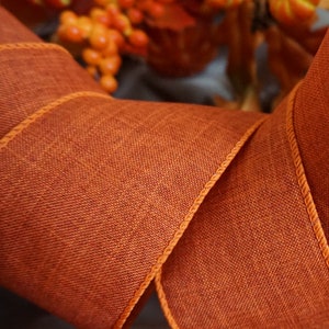 May include: Close-up of a roll of orange fabric ribbon with a textured weave and a decorative edge. The ribbon is likely used for crafting or decorating, possibly for autumn-themed projects.