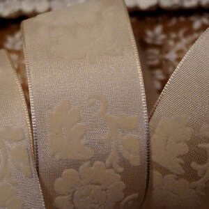 May include: White satin ribbon with an embossed floral pattern. The ribbon is folded in thirds, showing the pattern on both sides.