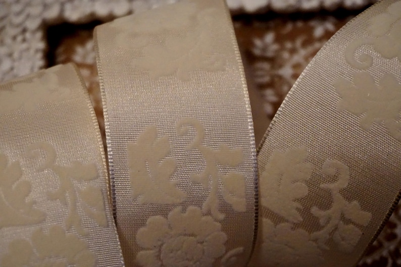 May include: White satin ribbon with an embossed floral pattern. The ribbon is folded in thirds, showing the pattern on both sides.