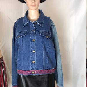 May include: A blue denim jacket with a red and blue embroidered trim around the bottom. The jacket has a button closure and two pockets. The model is wearing a black leather skirt and a black hat.