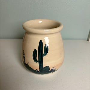 May include: A small, beige ceramic jar with a wide opening. The jar is decorated with a dark green cactus and desert landscape design. The jar is sitting on a white surface.