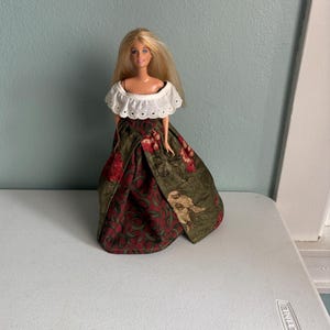 May include: A blonde doll wearing a white off-the-shoulder top and a long, patterned skirt. The skirt features a dark green background with red floral and bird designs. The doll is standing on a white surface.