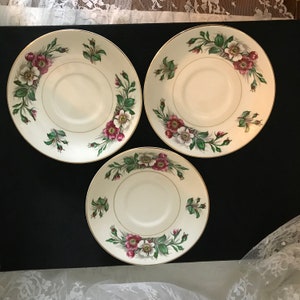 May include: Three white ceramic saucers with a gold rim and a floral design featuring pink and white flowers and green leaves.