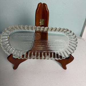 May include: A clear, oval-shaped glass serving dish with a ruffled edge. The dish is supported by a wooden display stand. The interior of the dish has a textured pattern. The background is a light blue wall.