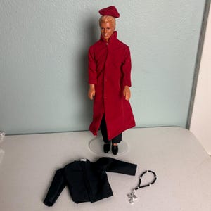Op de afbeelding: Een pop gekleed in een lange rode jas, zwarte broek en zwarte schoenen, met een bijpassende rode hoed. Een zwart jasje en een rozenkrans met een zilveren kruis liggen naast de pop. De pop heeft blond haar.
