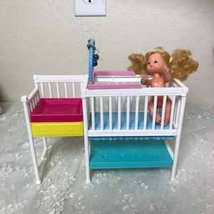 May include: A white plastic doll's cot with a pink and yellow changing table and a blue and white cot mattress. A doll with blonde hair is sitting in the cot.
