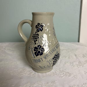 May include: A white ceramic pitcher with blue and black grapevine designs. The pitcher has a handle and the text "TRINKT WIE EURE VATER" is written on the side.