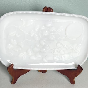 May include: A rectangular white serving dish with a floral design and scalloped edges. The dish has two decorative handles and is displayed on a wooden stand. The dish is approximately 12 inches long and 8 inches wide.