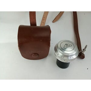 May include: A silver camera lens with the text "E. Leitz Wetzlar  Euflar  1:2,8  f=5cm  R" printed on the side. The lens is in a brown leather case with a strap.