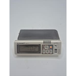 May include: A silver-toned Sonnet AG11 digital quartz travel alarm clock with a black folding case. The rectangular device features a digital display, a speaker grille, and several control buttons labeled Time, Alarm, Lock, and On/Off.