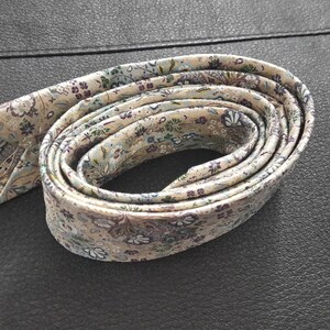 May include: A cream-colored patterned tie with a floral design in shades of purple, blue, and green. The tie is rolled up and tied in a knot.