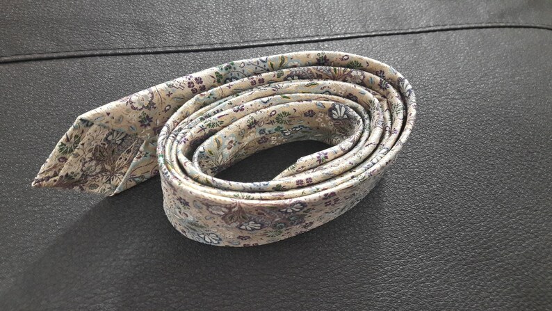 May include: A cream-colored patterned tie with a floral design in shades of purple, blue, and green. The tie is rolled up and tied in a knot.