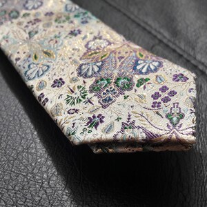 May include: A white tie with a floral pattern in shades of purple, green, and gold. The tie has a paisley design and is made of a silky fabric.