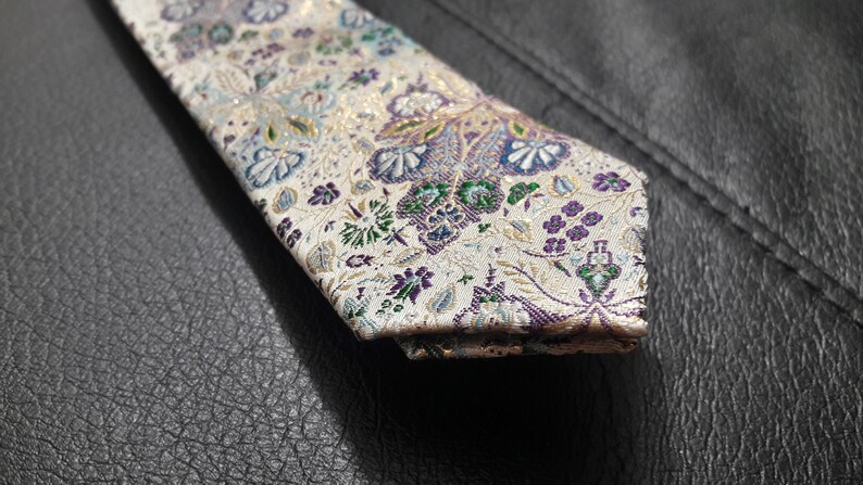 May include: A white tie with a floral pattern in shades of purple, green, and gold. The tie has a paisley design and is made of a silky fabric.