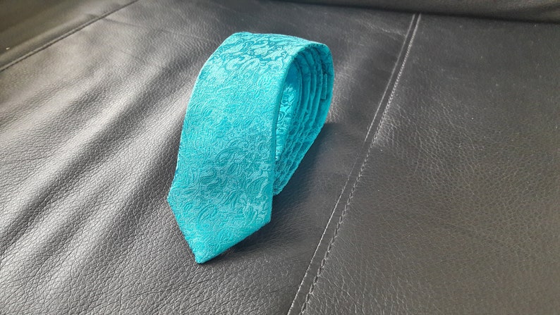 May include: A teal blue patterned tie with a floral design. The tie is rolled up and placed on a black leather surface.
