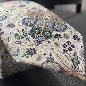 May include: A white tie with a floral pattern in shades of green, blue, and purple. The tie has a gold metallic thread woven into the fabric.