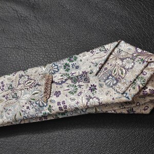 May include: A white tie with a floral pattern in shades of purple, green, and silver. The tie has a brown label with the word "Goode" printed on it.