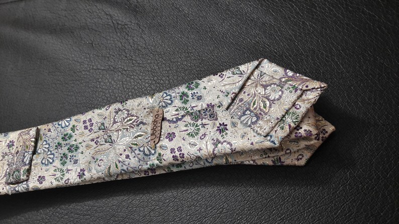 May include: A white tie with a floral pattern in shades of purple, green, and silver. The tie has a brown label with the word "Goode" printed on it.